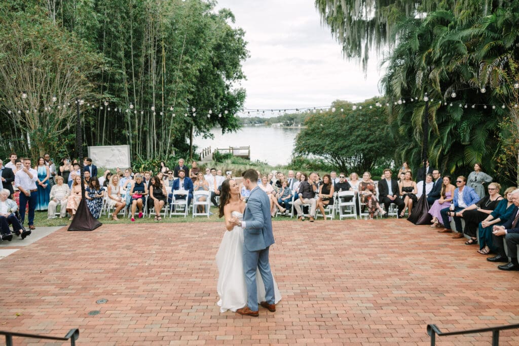 Wedding Venues Winter Park | Jennifer Holly Light and Airy Wedding Photographer in Orlando | A couple dances joyfully on a brick patio, surrounded by seated guests. Background features lush greenery, a lake, and string lights, creating a serene wedding scene.