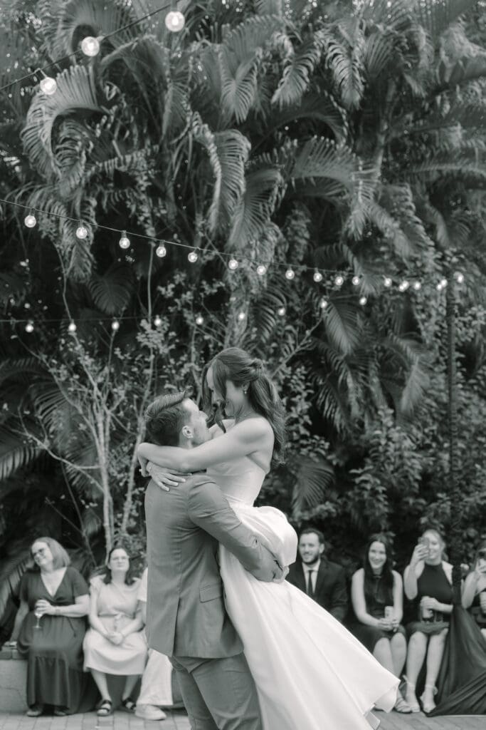 Outdoor Wedding Venues Orlando | Jennifer Holly Light and Airy Wedding Photographer in Orlando | A couple shares a joyful embrace during a wedding dance under string lights. Guests watch happily, surrounded by lush greenery. The mood is romantic and festive.