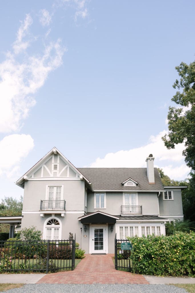 Garden Wedding Orlando | Jennifer Holly Light and Airy Wedding Photographer in Orlando | Elegant two-story gray house with white trim, surrounded by lush greenery. Features a brick path, iron fence, and chimney under a clear blue sky.