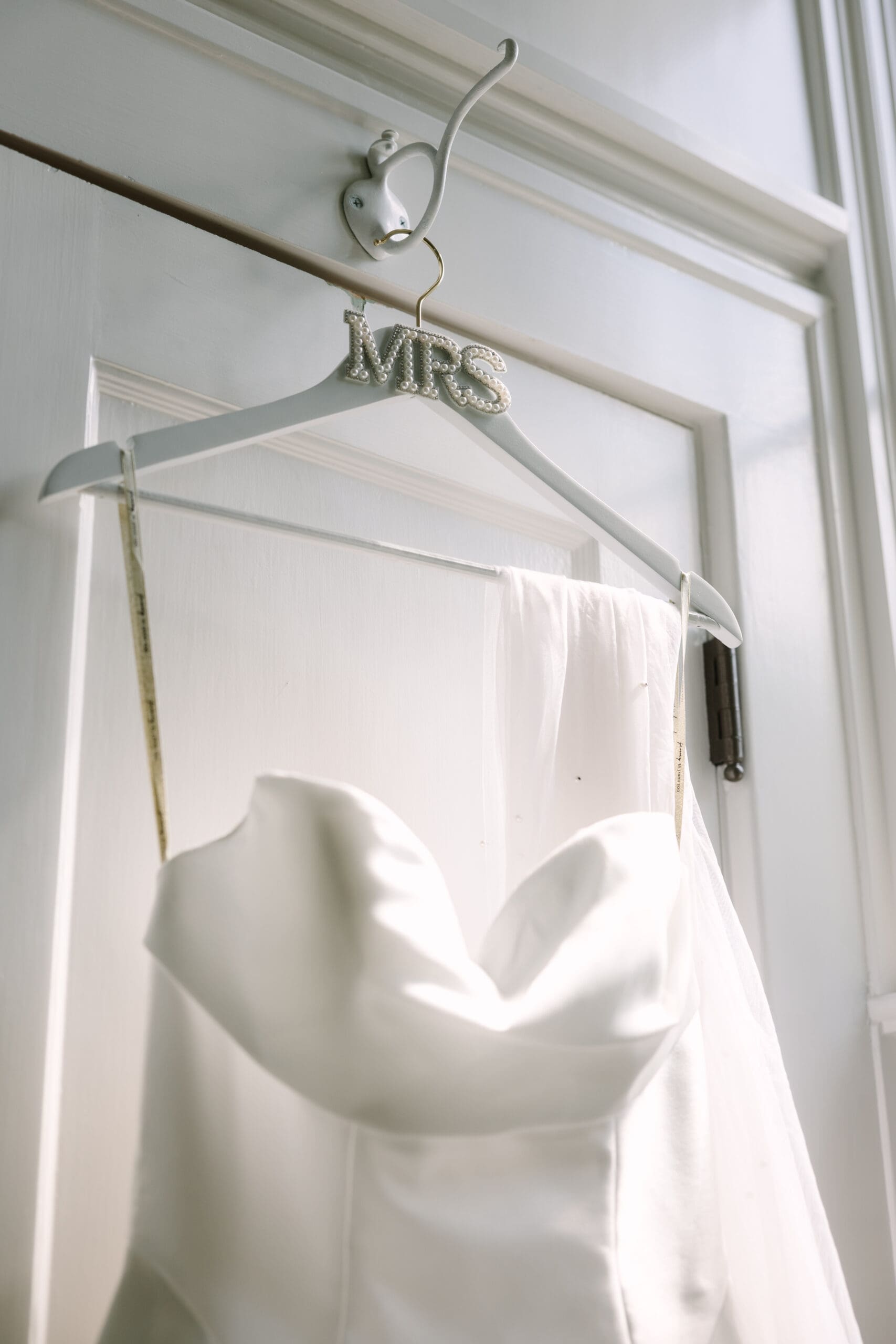 Winter Park Wedding Chapel | Jennifer Holly Light and Airy Wedding Photographer in Orlando | A white wedding dress with a strapless neckline hangs on a pearl-studded hanger labeled "MRS," set against a wooden door, evoking elegance.