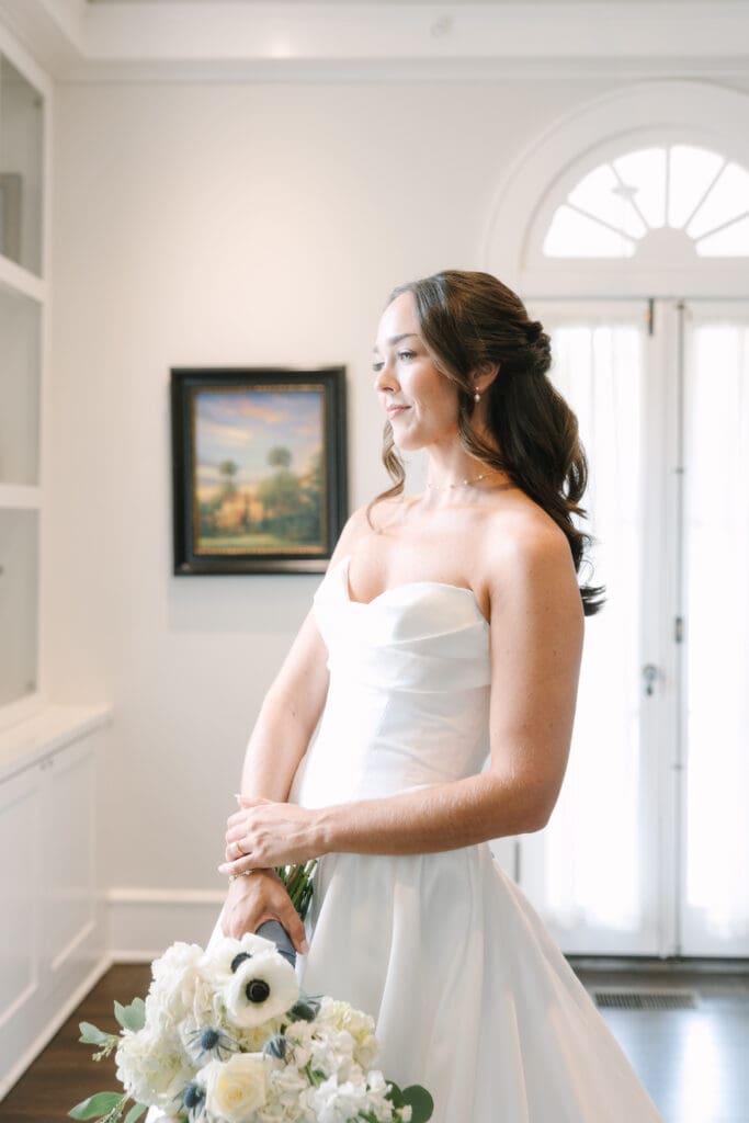 Wedding Venues Winter Park | Jennifer Holly Light and Airy Wedding Photographer in Orlando | A bride in a white strapless gown holds a bouquet with pale flowers. She stands in a bright room with an elegant archway and framed painting, looking serene.