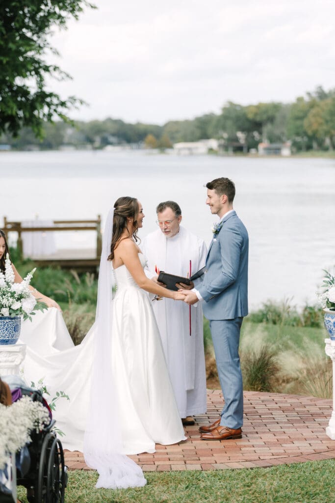 Outdoor Wedding Venues Orlando | Jennifer Holly Light and Airy Wedding Photographer in Orlando | A bride in a white gown and a groom in a blue suit exchange vows by a lakeside. A priest officiates, creating a serene and joyful atmosphere.