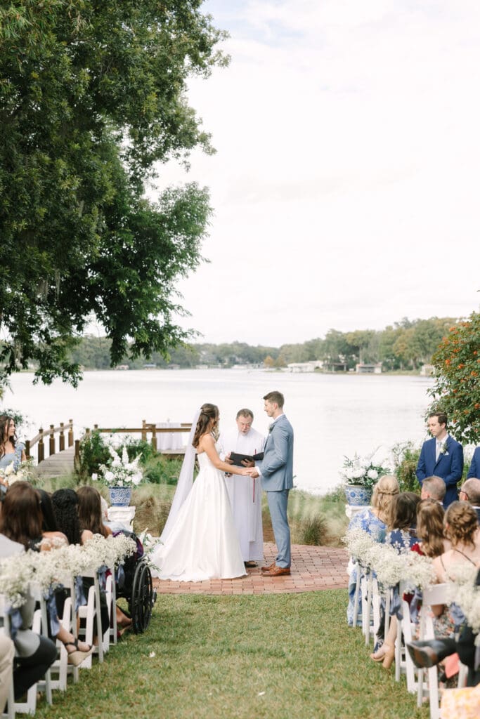 Winter Park Wedding | Jennifer Holly Light and Airy Wedding Photographer in Orlando | A bride and groom stand hand in hand during an outdoor wedding by a serene lake, with a priest officiating. Guests sit on white chairs adorned with flowers.