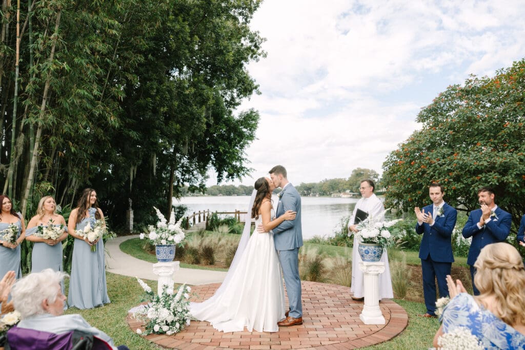 Garden Wedding Venues in Orlando | Jennifer Holly Light and Airy Wedding Photographer in Orlando | A bride and groom share a kiss during an outdoor wedding by a lake. The bride wears a white gown, and the groom a light blue suit. A clergyman looks on, while lush greenery and floral arrangements create a serene atmosphere.
