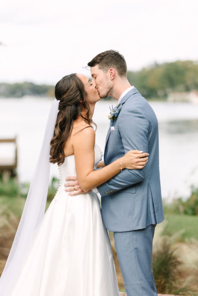Wedding Venues Winter Park | Jennifer Holly Light and Airy Wedding Photographer in Orlando | A bride in a white dress and veil kisses a groom in a blue suit by a lakeside. The scene is romantic and serene, conveying joy and love.