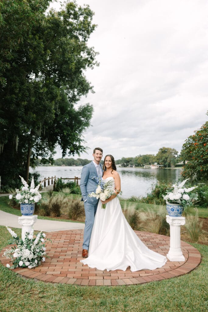 Wedding Venues Winter Park | Jennifer Holly Light and Airy Wedding Photographer in Orlando | A couple stands together on a brick circle, smiling in formal wedding attire. They're by a lakeside with floral arrangements and lush greenery around them.