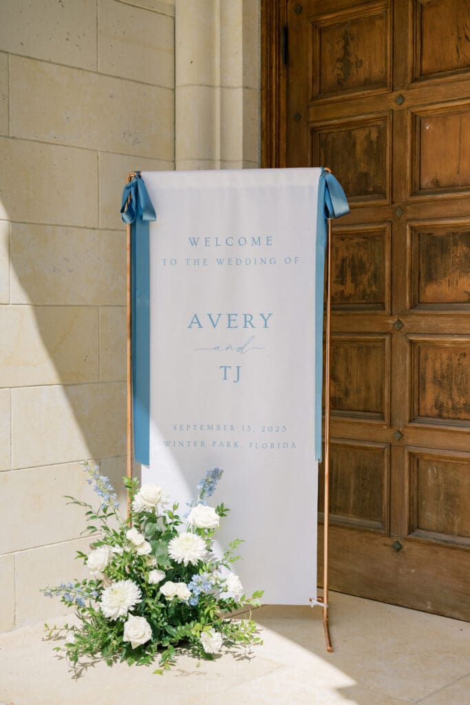 Garden Wedding Venues in Orlando | Jennifer Holly Light and Airy Wedding Photographer in Orlando | Wedding sign outside wooden door reads "Welcome to the Wedding of Avery and TJ." It features blue ribbons, and white flowers with greenery are arranged below.