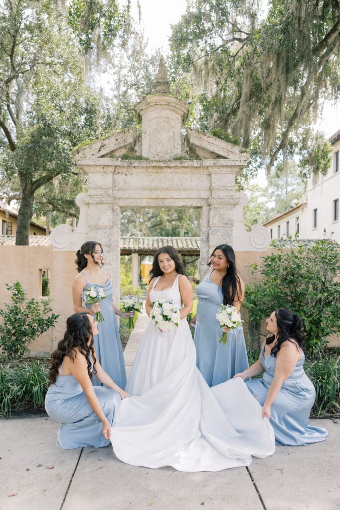Wedding Venues Winter Park | Jennifer Holly Light and Airy Wedding Photographer in Orlando | Bride in white stands joyfully with four bridesmaids in blue dresses. They're under a stone arch, surrounded by greenery and trees, creating a serene atmosphere.