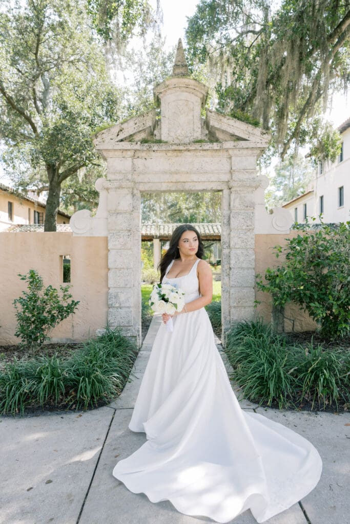 Outdoor Wedding Venues Orlando | Jennifer Holly Light and Airy Wedding Photographer in Orlando | Bride in a flowing white gown holding a bouquet, stands under an arch in a lush garden setting. The scene is serene with soft sunlight filtering through trees.