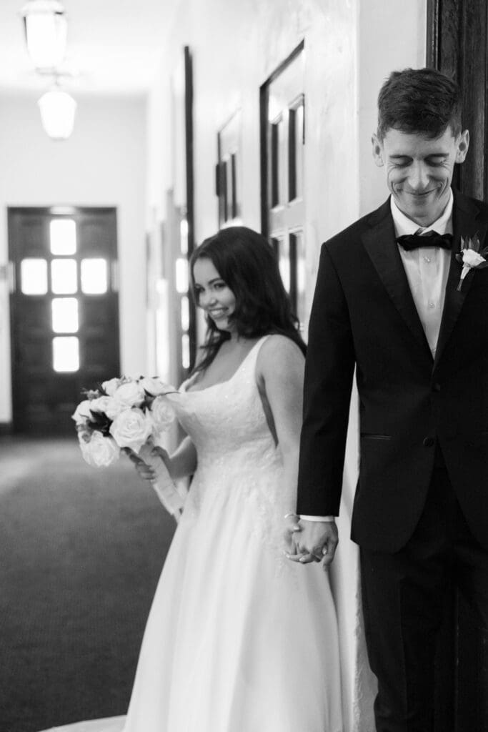 Winter Park Wedding Chapel | Jennifer Holly Light and Airy Wedding Photographer in Orlando | A bride and groom stand holding hands around a corner, not seeing each other. The bride smiles with a bouquet, and the groom wears a shy smile. Black and white.