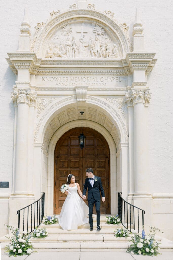 Outdoor Wedding Venues Orlando | Jennifer Holly Light and Airy Wedding Photographer in Orlando | A bride and groom hold hands, smiling as they exit a chapel with ornate carvings above the wooden door. Flowers adorn the steps. Joyful, elegant scene.