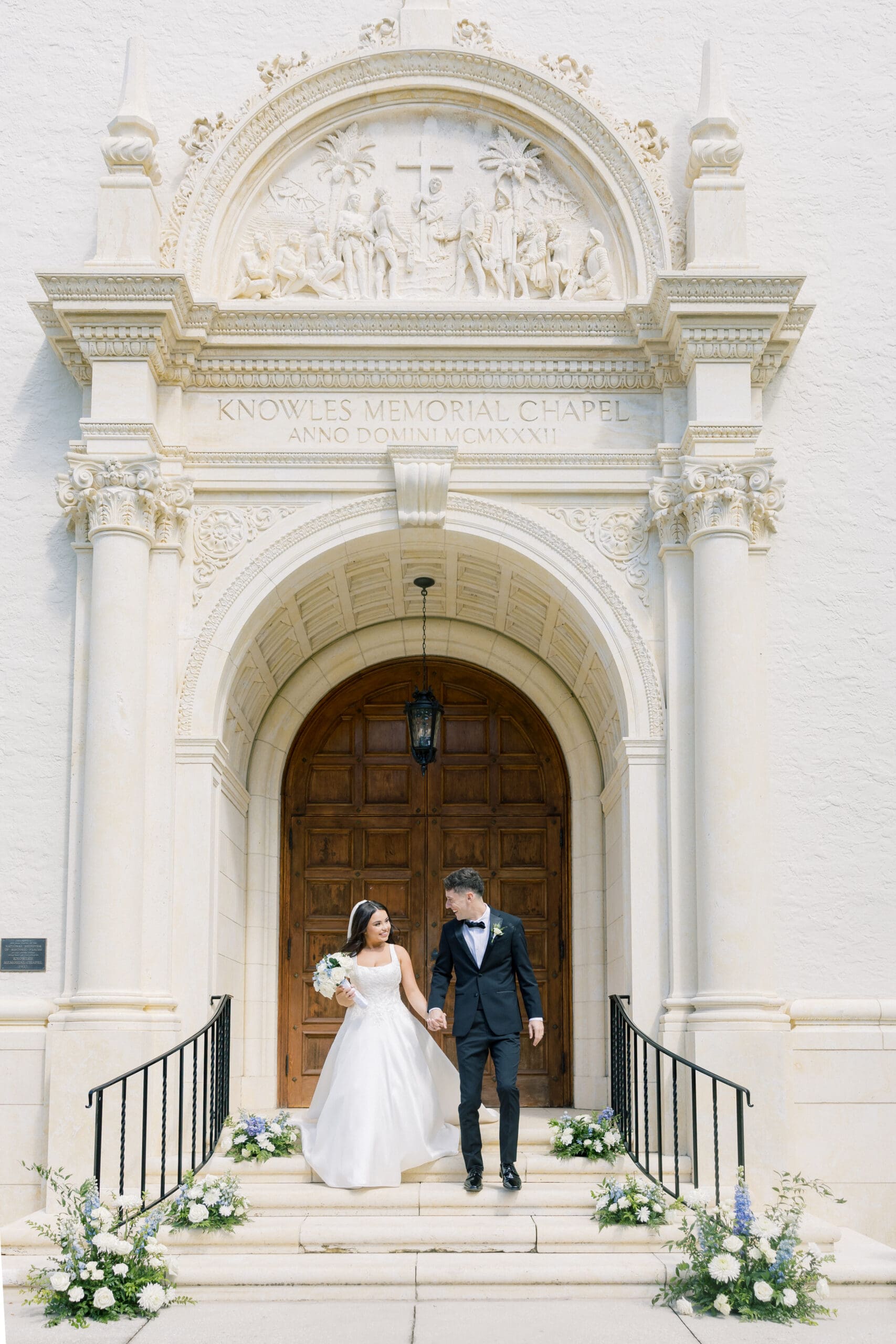 Outdoor Wedding Venues Orlando | Jennifer Holly Light and Airy Wedding Photographer in Orlando | A bride and groom hold hands, smiling as they exit a chapel with ornate carvings above the wooden door. Flowers adorn the steps. Joyful, elegant scene.