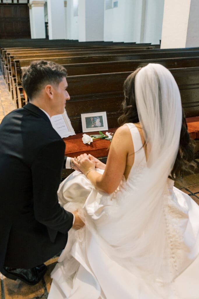 Winter Park Wedding Chapel | Jennifer Holly Light and Airy Wedding Photographer in Orlando | A bride and groom kneel in a church, holding hands. In front of them is a bench with a small framed photo, flowers, and paper. The moment feels intimate.