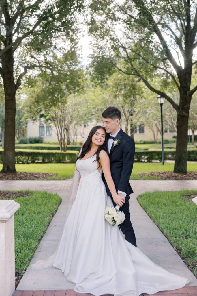 Garden Wedding Venues in Orlando | Jennifer Holly Light and Airy Wedding Photographer in Orlando | A bride in a flowing white gown and a groom in a black suit pose romantically in a sunlit garden path, surrounded by lush green trees.