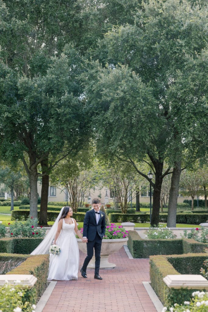 Wedding Venues Winter Park | Jennifer Holly Light and Airy Wedding Photographer in Orlando | A bride in a white dress and groom in a dark suit walk hand in hand through a manicured garden. Tall trees and vibrant flowers create a serene backdrop.