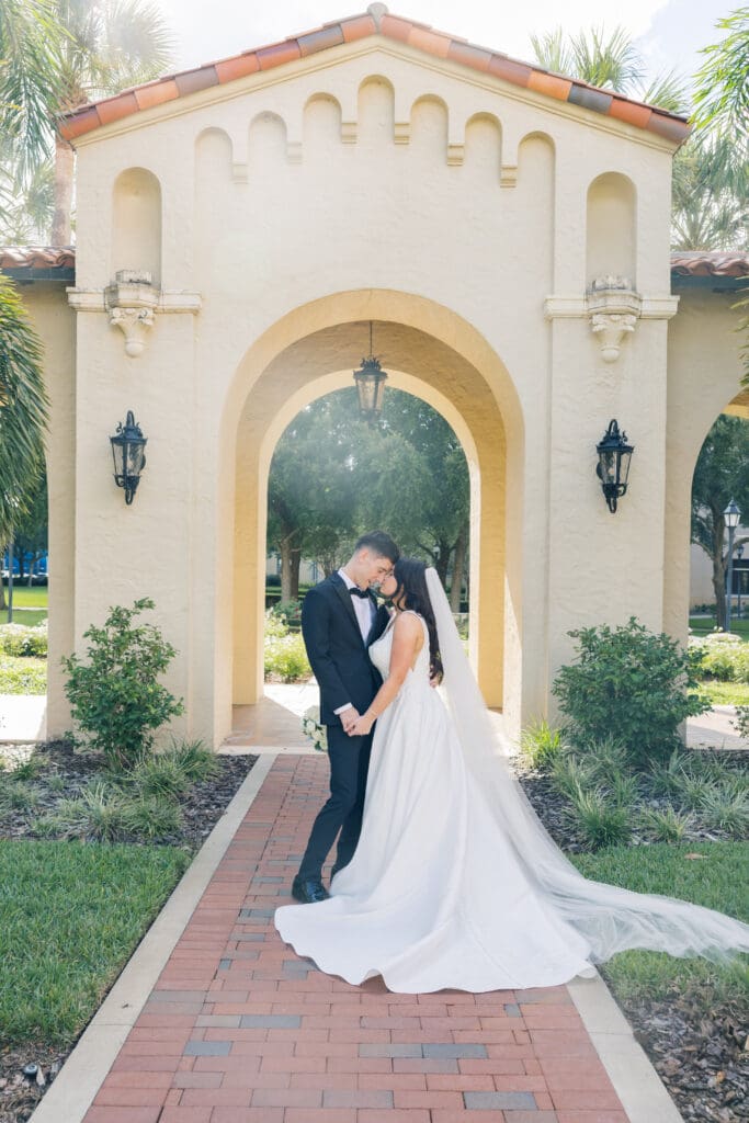 Outdoor Wedding Venues Orlando | Jennifer Holly Light and Airy Wedding Photographer in Orlando | A bride and groom embrace in front of a sunlit, arched building with lush greenery. The bride's white gown and veil trail elegantly, conveying romance.