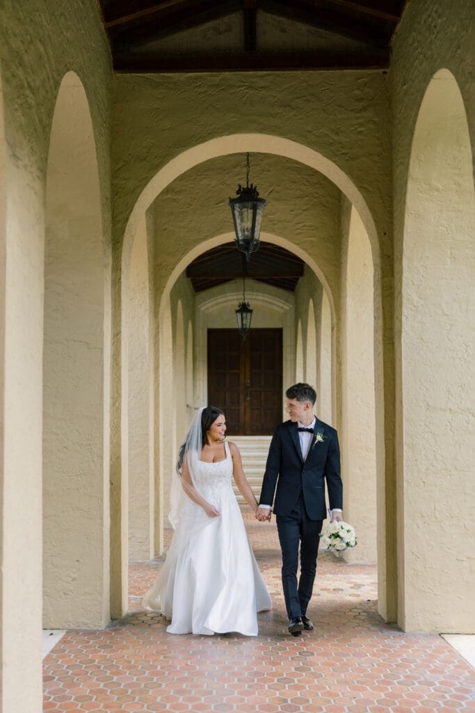 Winter Park Wedding Chapel | Jennifer Holly Light and Airy Wedding Photographer in Orlando | Bride in white gown and groom in black suit holding hands, walking down an elegant, arched hallway. They smile at each other, conveying happiness.