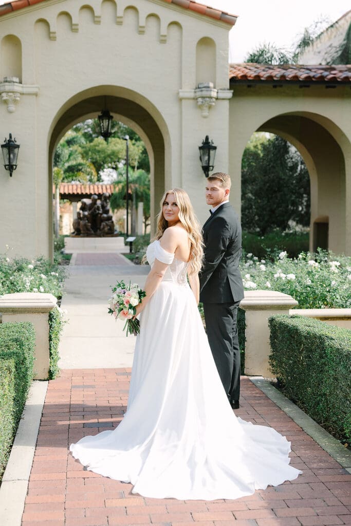 Alfond Inn Winter Park | Jennifer Holly Light and Airy Wedding Photographer in Orlando | A bride in a white gown holding a bouquet and a groom in a black suit stand on a brick path, smiling, in front of an elegant archway and greenery.