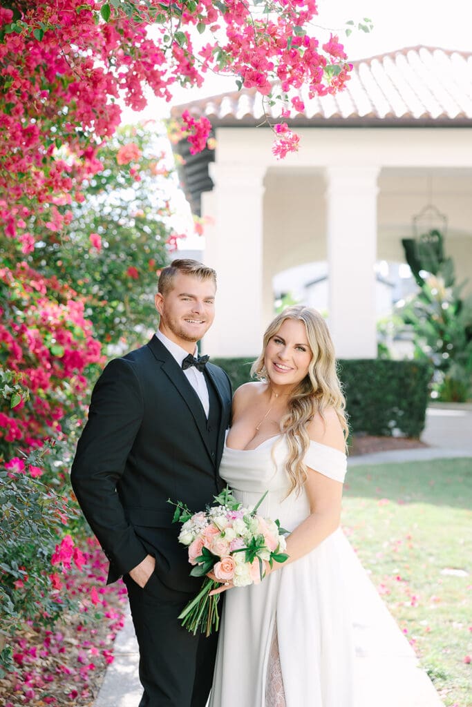 Alfond Inn Winter Park | Jennifer Holly Light and Airy Wedding Photographer in Orlando | A couple in wedding attire smiling under vibrant pink bougainvillea, with a sunlit garden and elegant building in the background conveying joy and romance.