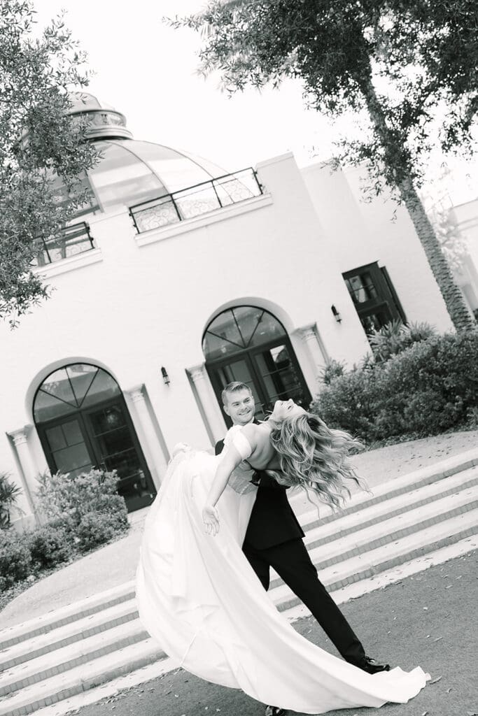 Alfond Inn Winter Park | Jennifer Holly Light and Airy Wedding Photographer in Orlando | A black-and-white photo of a man playfully lifting a woman in a wedding dress in front of a grand building with arched windows. Joyful and romantic atmosphere.