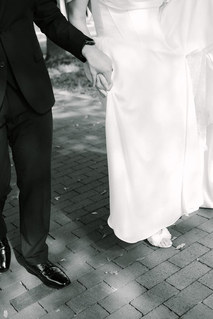Alfond Inn Winter Park | Jennifer Holly Light and Airy Wedding Photographer in Orlando | A couple holding hands, walking on a brick path. The man is in a dark suit, the woman in a white flowing dress. It's a black-and-white image.