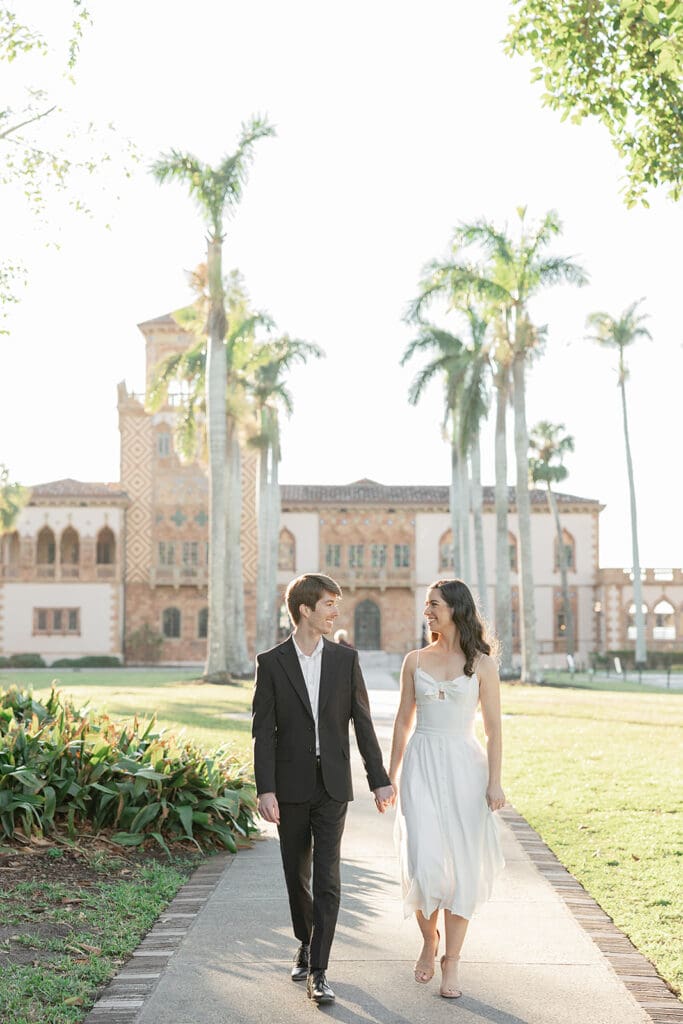 The Ringling Engagement Photos | Jennifer Holly Light and Airy Wedding Photographer in Orlando | A couple walks hand in hand on a sunny path, smiling at each other. They're in formal attire, with a historic building and tall palm trees in the background.