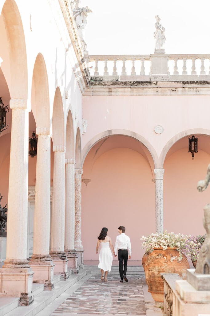 The Ringling Engagement Photos | Jennifer Holly Light and Airy Wedding Photographer in Orlando | A couple holds hands, walking along a sunlit courtyard with peach-colored walls and arched columns. Floral pots add a romantic, serene atmosphere.