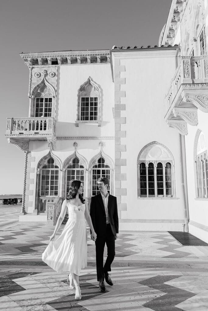 The Ringling Engagement Photos | Jennifer Holly Light and Airy Wedding Photographer in Orlando | A couple walks hand in hand in front of an ornate building with arched windows. The black and white image conveys elegance and romance.