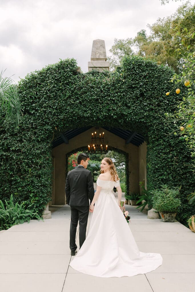 Sydonie Mansion Wedding | Jennifer Holly Light and Airy Wedding Photographer in Orlando | A bride in a flowing white dress and a groom in a black suit walk hand in hand. They face an ivy-covered archway, conveying romance and intimacy.