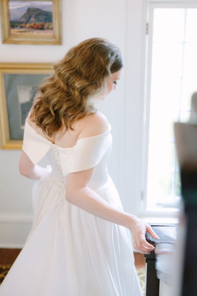 Sydonie Mansion Wedding | Jennifer Holly Light and Airy Wedding Photographer in Orlando | Bride in elegant, off-the-shoulder white gown stands near a window, looking down. Soft lighting and delicate curls create a serene, romantic mood.