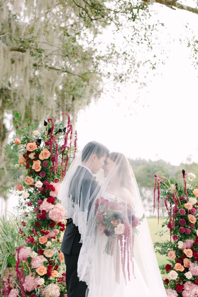 Sydonie Mansion Wedding | Jennifer Holly Light and Airy Wedding Photographer in Orlando | A bride and groom embrace under a veil, surrounded by lush floral arches of red, peach, and pink roses, set against a serene outdoor backdrop. Romantic and tender atmosphere.