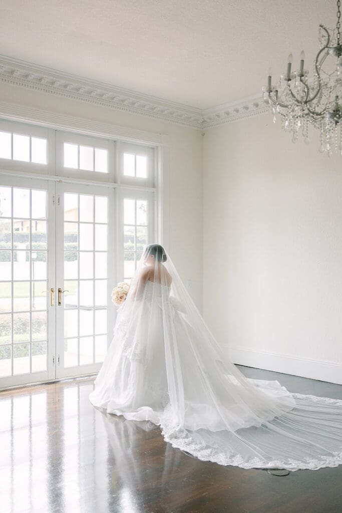 Wedding Venues in Central Florida | Jennifer Holly Light and Airy Wedding Photographer in Orlando | A bride in a flowing white gown and veil stands by large windows, holding a bouquet. The room is elegant with a chandelier and hardwood floors.
