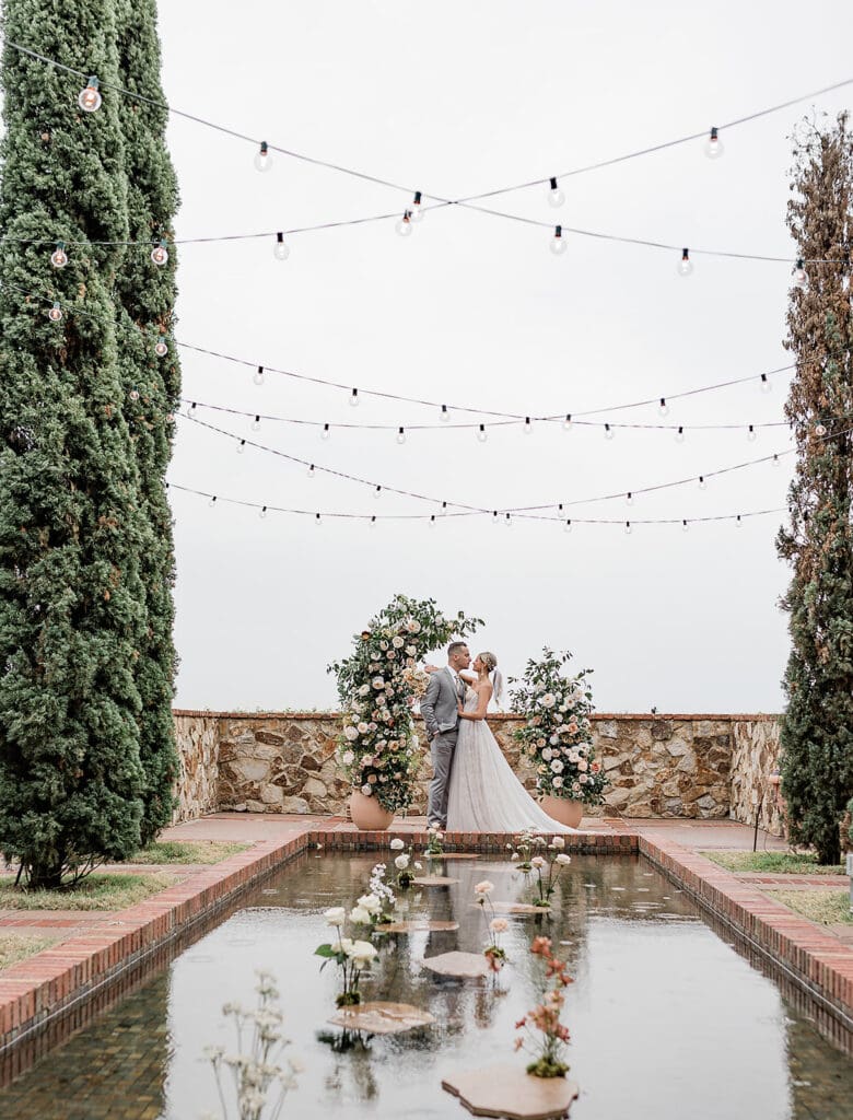 Wedding Venues in Central Florida | Jennifer Holly Light and Airy Wedding Photographer in Orlando | A couple embraces in a garden setting with string lights overhead. They're framed by floral arrangements next to a reflective pool. Romantic atmosphere.