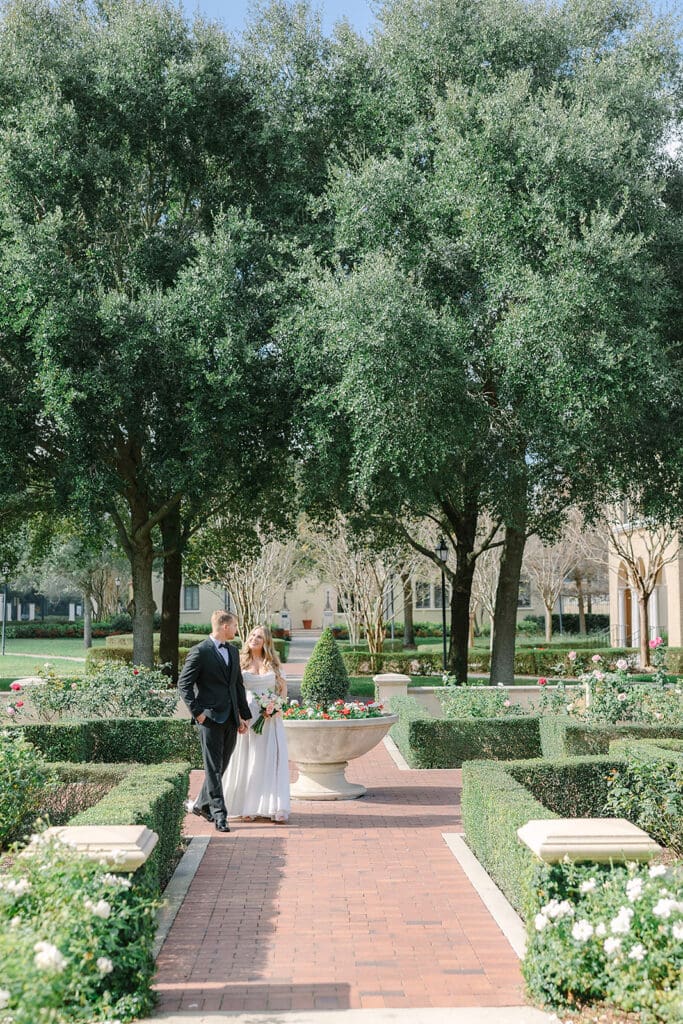 Wedding Venues in Central Florida | Jennifer Holly Light and Airy Wedding Photographer in Orlando | A couple walks along a brick pathway in a lush garden, surrounded by manicured hedges and large trees. The mood is serene and romantic.
