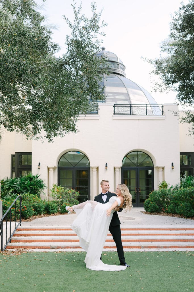 Wedding Venues in Central Florida | Jennifer Holly Light and Airy Wedding Photographer in Orlando | A groom in a tuxedo joyfully lifts his bride, wearing a flowing white gown, in front of a grand building with arched windows and greenery.