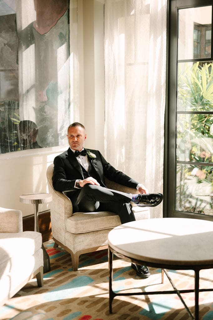 Casa Feliz Wedding | Jennifer Holly Light and Airy Wedding Photographer in Orlando | A man in a tuxedo sits confidently on an armchair in a sunlit room. He appears relaxed, surrounded by soft furnishings and natural light filtering through a window.