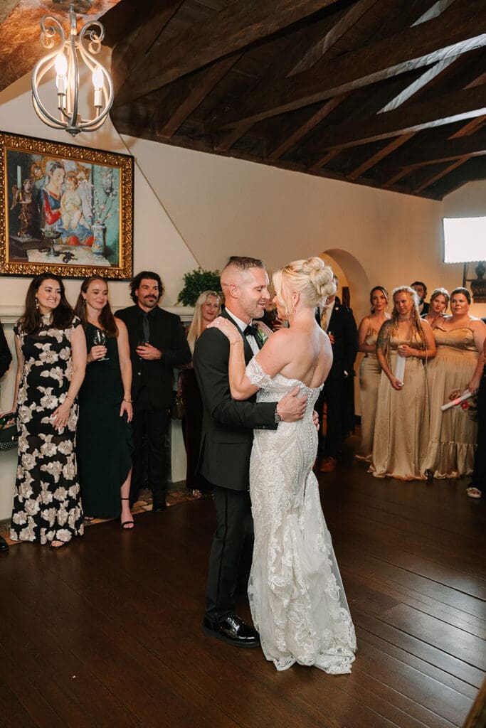 Casa Feliz Wedding | Jennifer Holly Light and Airy Wedding Photographer in Orlando | A couple shares a joyful first dance at their wedding, surrounded by smiling guests in elegant attire in a warmly lit room with wooden beams.