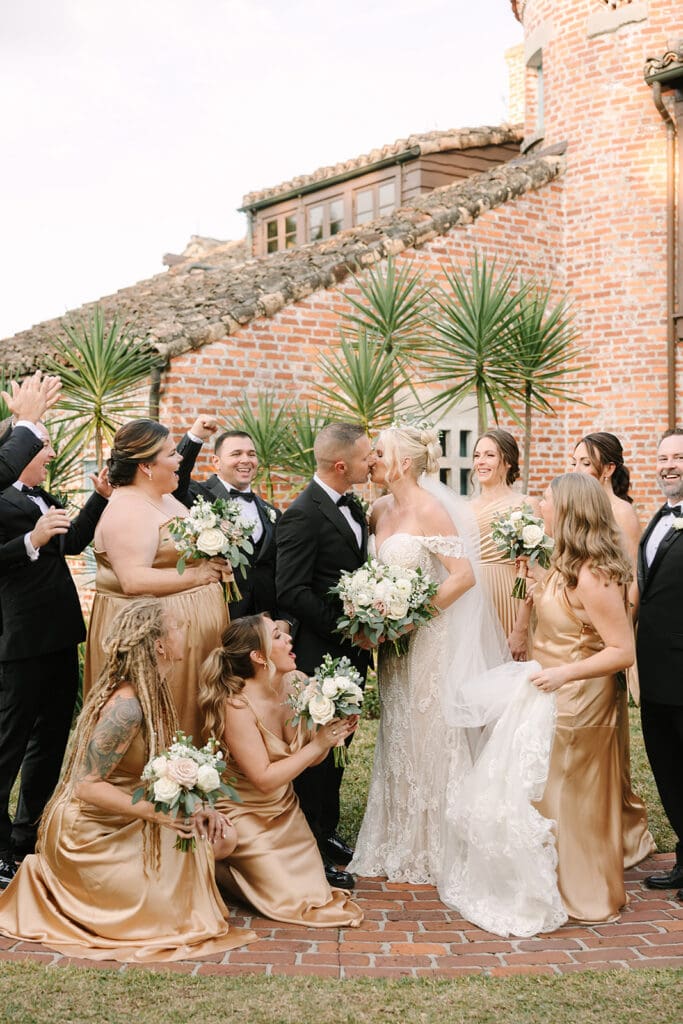 Casa Feliz Wedding | Jennifer Holly Light and Airy Wedding Photographer in Orlando | Wedding party celebrating with joy as bride and groom kiss. Bride in lace gown, bridesmaids in gold dresses, groomsmen in black suits, against a brick building.