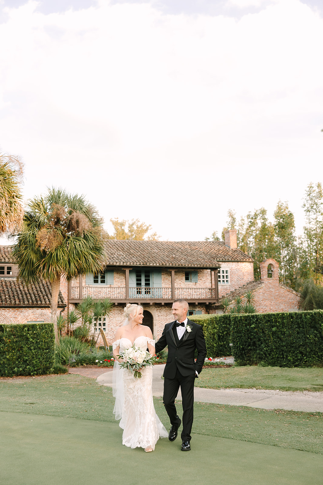 Casa Feliz Wedding | Jennifer Holly Light and Airy Wedding Photographer in Orlando | A bride and groom walk arm in arm on a green lawn, smiling at each other. Behind them is a rustic brick house with palm trees and hedges, under a soft sky.