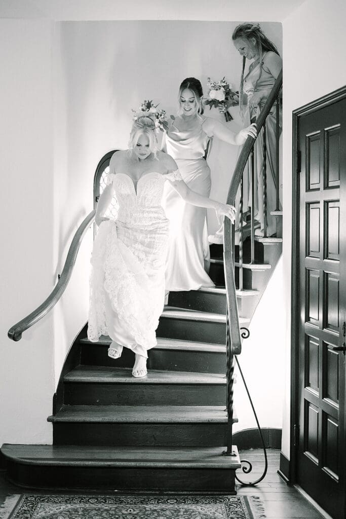 Casa Feliz Wedding | Jennifer Holly Light and Airy Wedding Photographer in Orlando | Bride in a lace gown descends a wooden spiral staircase, flanked by two bridesmaids holding bouquets. The mood is joyful and elegant.