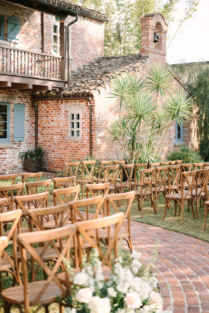 Casa Feliz Wedding | Jennifer Holly Light and Airy Wedding Photographer in Orlando | Outdoor wedding setup with wooden chairs arranged on a brick patio, beside a charming brick house with blue shutters and lush greenery, creating a serene atmosphere.