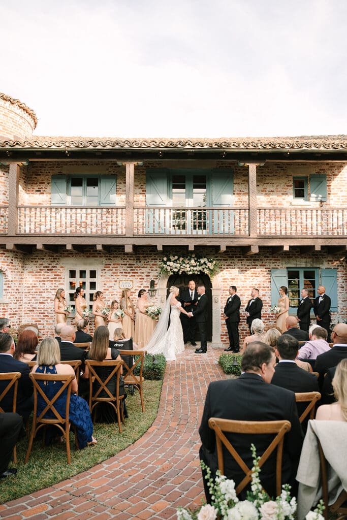 Casa Feliz Wedding | Jennifer Holly Light and Airy Wedding Photographer in Orlando | Wedding ceremony outside a rustic brick building with teal shutters. Bride and groom hold hands under a flower arch, surrounded by bridal party and seated guests.