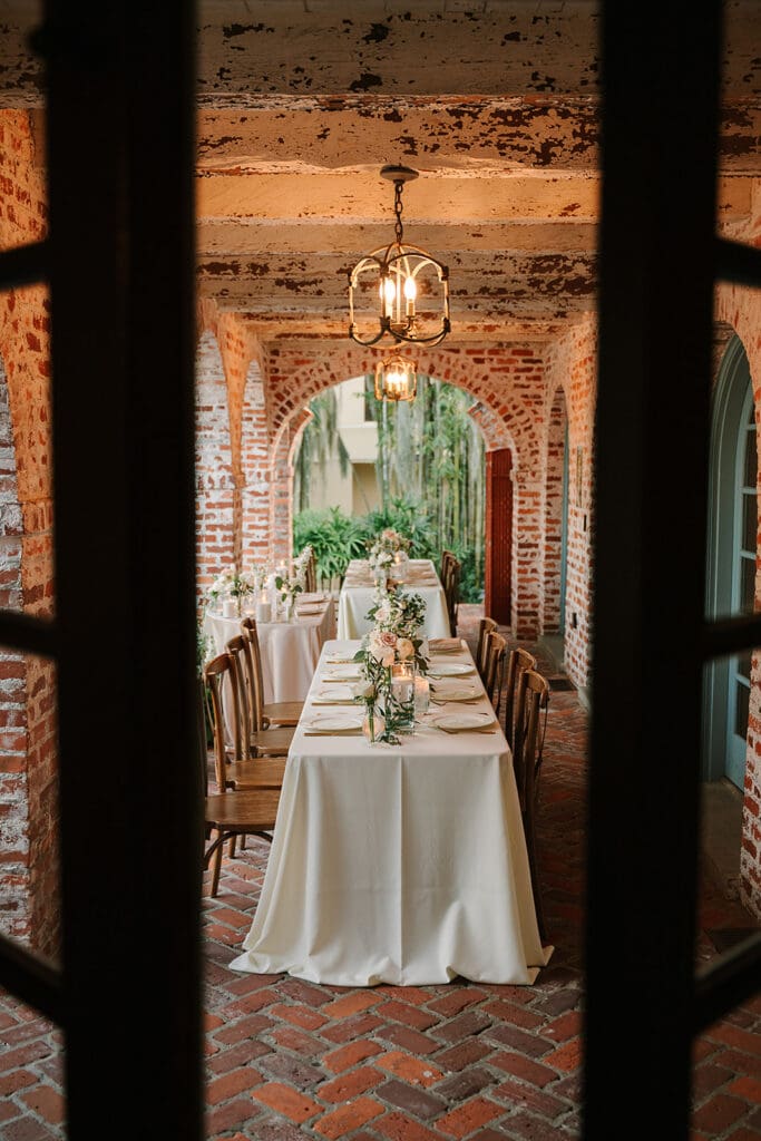 Casa Feliz Wedding | Jennifer Holly Light and Airy Wedding Photographer in Orlando | A rustic outdoor dining setting with long, elegant tables covered in white cloth, adorned with foliage, lit candles, and simple, elegant tableware. The scene is framed by brick arches, evoking a warm, inviting atmosphere.