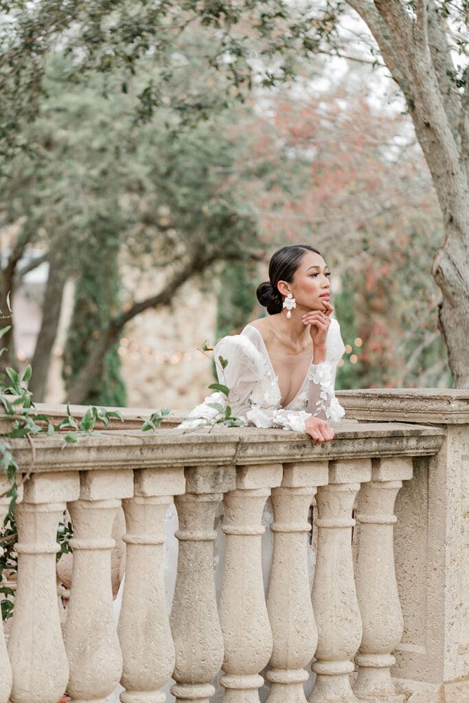 Wedding Venues in Central Florida | Jennifer Holly Light and Airy Wedding Photographer in Orlando | A woman in a white dress leans thoughtfully on a stone balustrade, surrounded by trees with a blurred, serene garden backdrop.