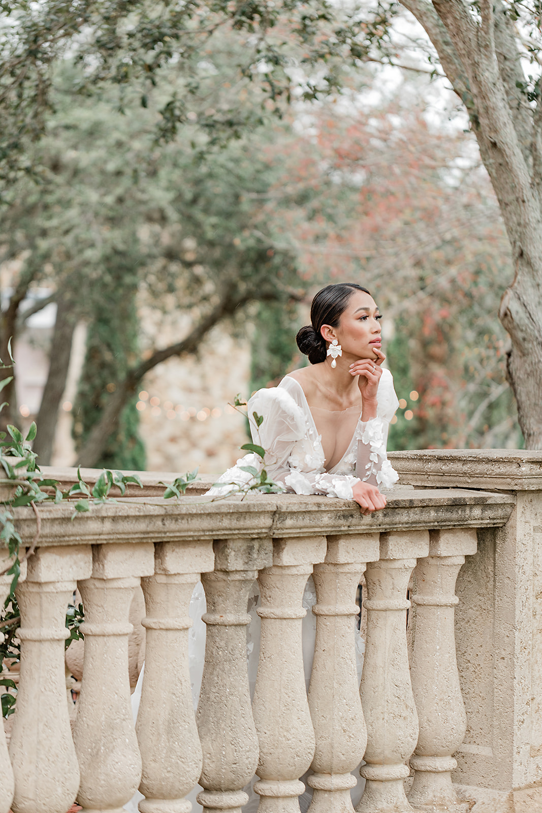 Wedding Venues in Central Florida | Jennifer Holly Light and Airy Wedding Photographer in Orlando | A woman in a white dress leans thoughtfully on a stone balustrade, surrounded by trees with a blurred, serene garden backdrop.