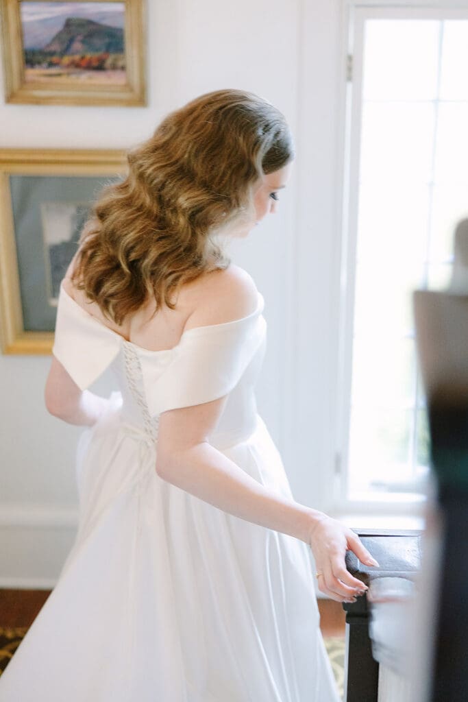 Sydonie Mansion Wedding Photos | Jennifer Holly Light and Airy Wedding Photographer in Orlando | Bride in off-the-shoulder white gown gracefully descends stairs, her wavy hair cascading. Soft light floods through a window, creating a serene atmosphere.