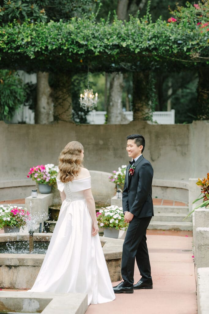 Sydonie Mansion Wedding Photos | Jennifer Holly Light and Airy Wedding Photographer in Orlando | Bride and groom smiling at each other in a garden setting. She wears a white gown; he wears a black suit. Surrounding them are blooming flowers.