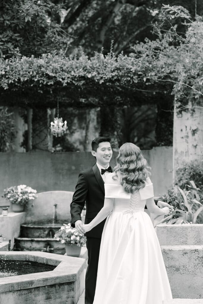 Sydonie Mansion Wedding Photos | Jennifer Holly Light and Airy Wedding Photographer in Orlando | A couple is elegantly dressed for a wedding. The groom in a tuxedo smiles at the bride in a white gown. They're near a garden fountain, creating a romantic atmosphere.