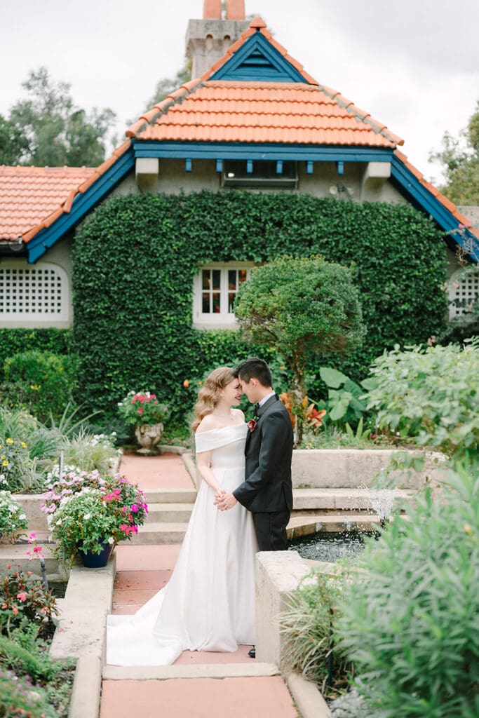 Sydonie Mansion Wedding Photos | Jennifer Holly Light and Airy Wedding Photographer in Orlando | A couple stands closely, sharing an intimate moment in a garden. They are in front of a charming ivy-covered house with a red-tiled roof, surrounded by colorful flowers.