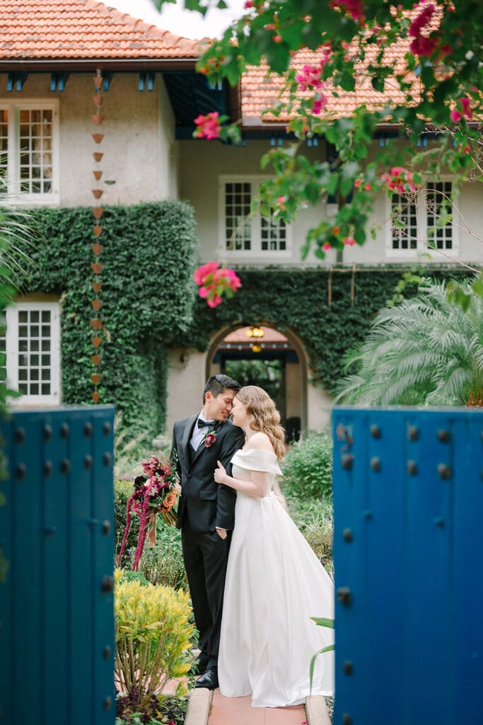Sydonie Mansion Wedding Photos | Jennifer Holly Light and Airy Wedding Photographer in Orlando | A bride in a flowing white gown embraces a groom in a black suit in a garden. They're framed by open blue gates and a charming ivy-covered house.